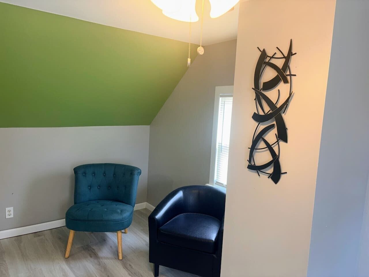 A room with a sloped ceiling, a green wall, two chairs, and a wall-mounted abstract art piece.