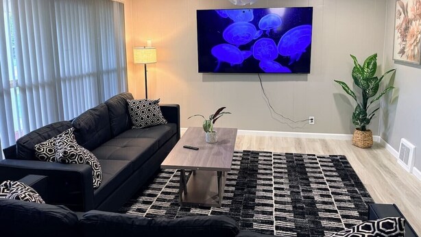 Ein modernes Wohnzimmer mit einem schwarzen Sofa, einem Couchtisch, einem großen Flachbildfernseher, einem Deckenventilator und einem gemusterten Teppich.