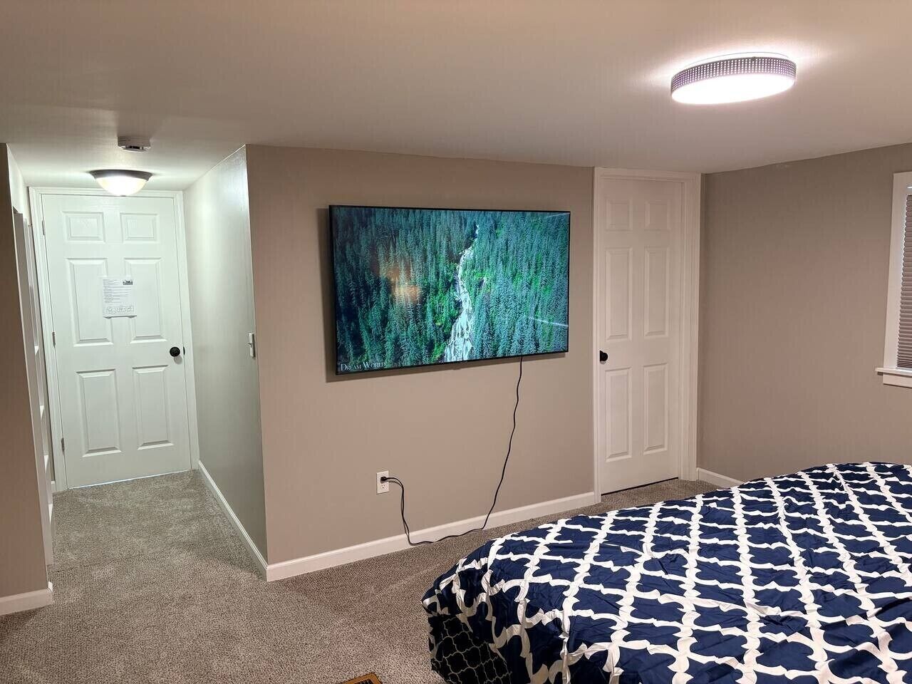 Ein Schlafzimmer mit einem Bett, einem an der Wand befestigten Fernseher und einer Tür zu einem anderen Raum.