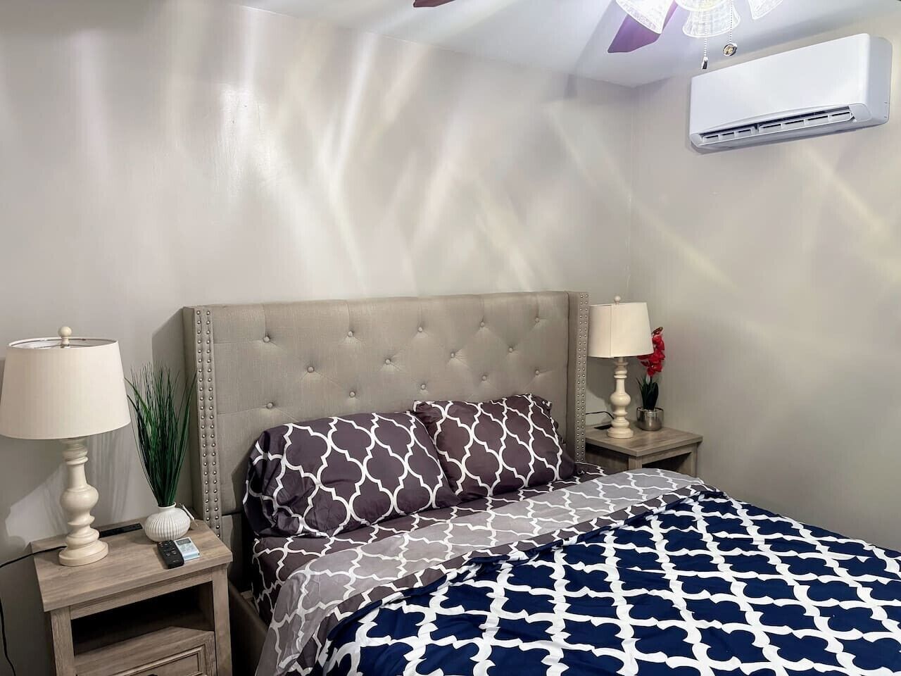 A bedroom with a bed, two bedside tables, lamps, a wall-mounted air conditioner, and a potted plant.