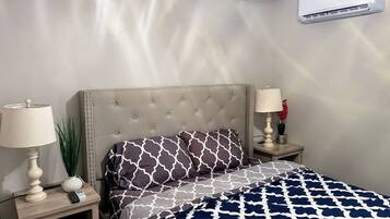 A bedroom with a bed, two bedside tables, lamps, a wall-mounted air conditioner, and a potted plant.