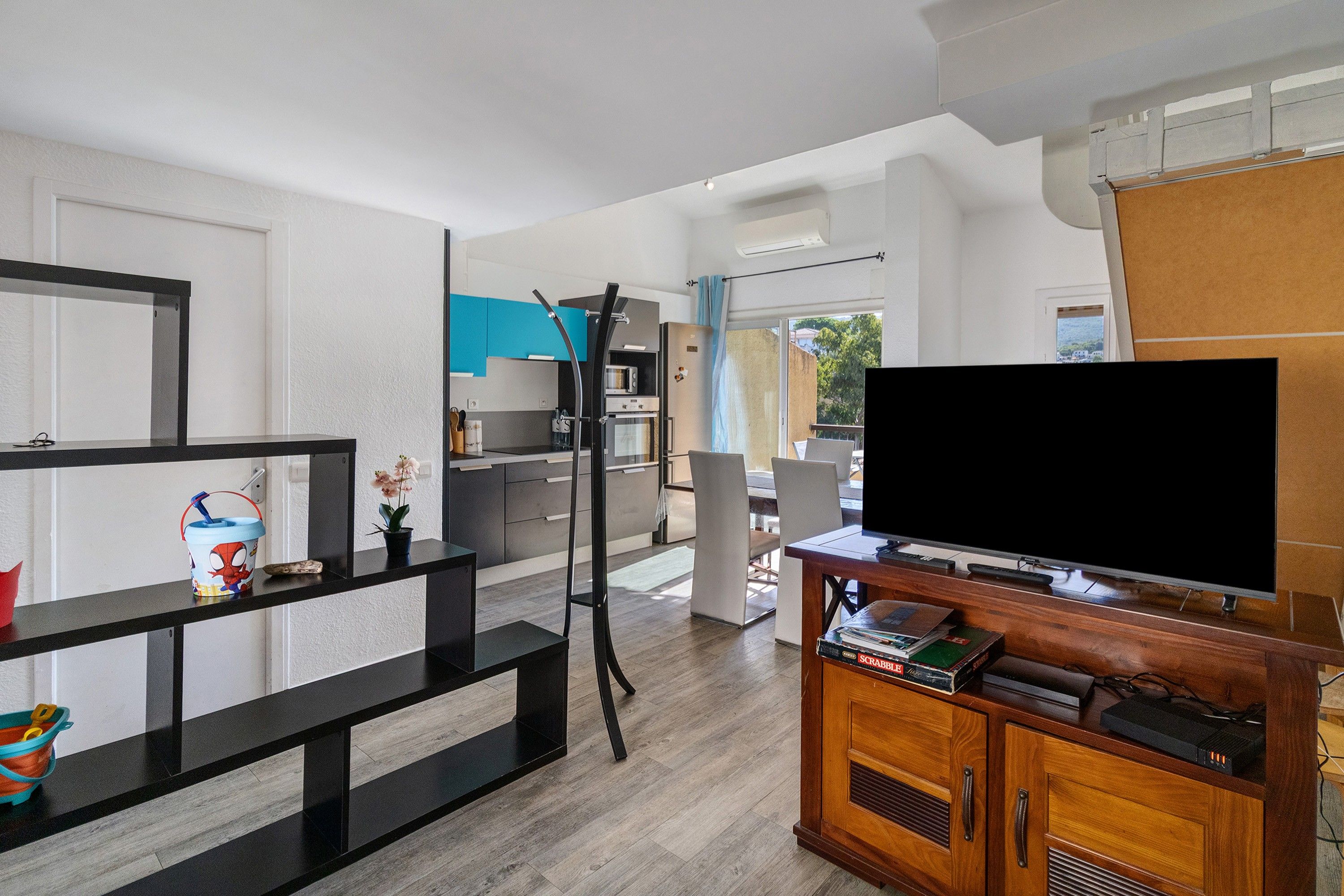 Un salon moderne avec un téléviseur à écran plat, un meuble de rangement en bois et, au fond, un coin cuisine.