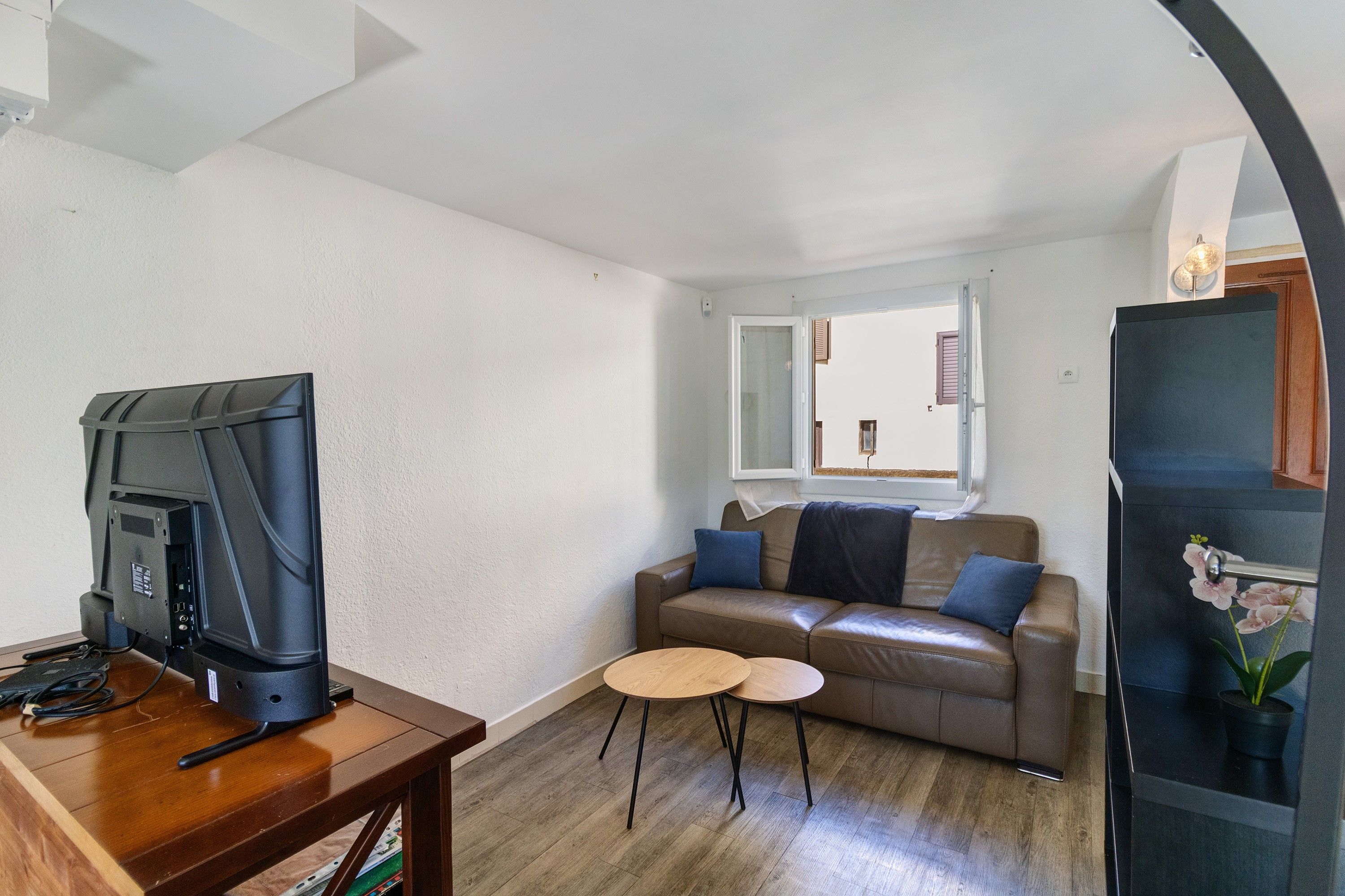 A living room with a television, a brown leather sofa, two small round tables, and a window with blinds.