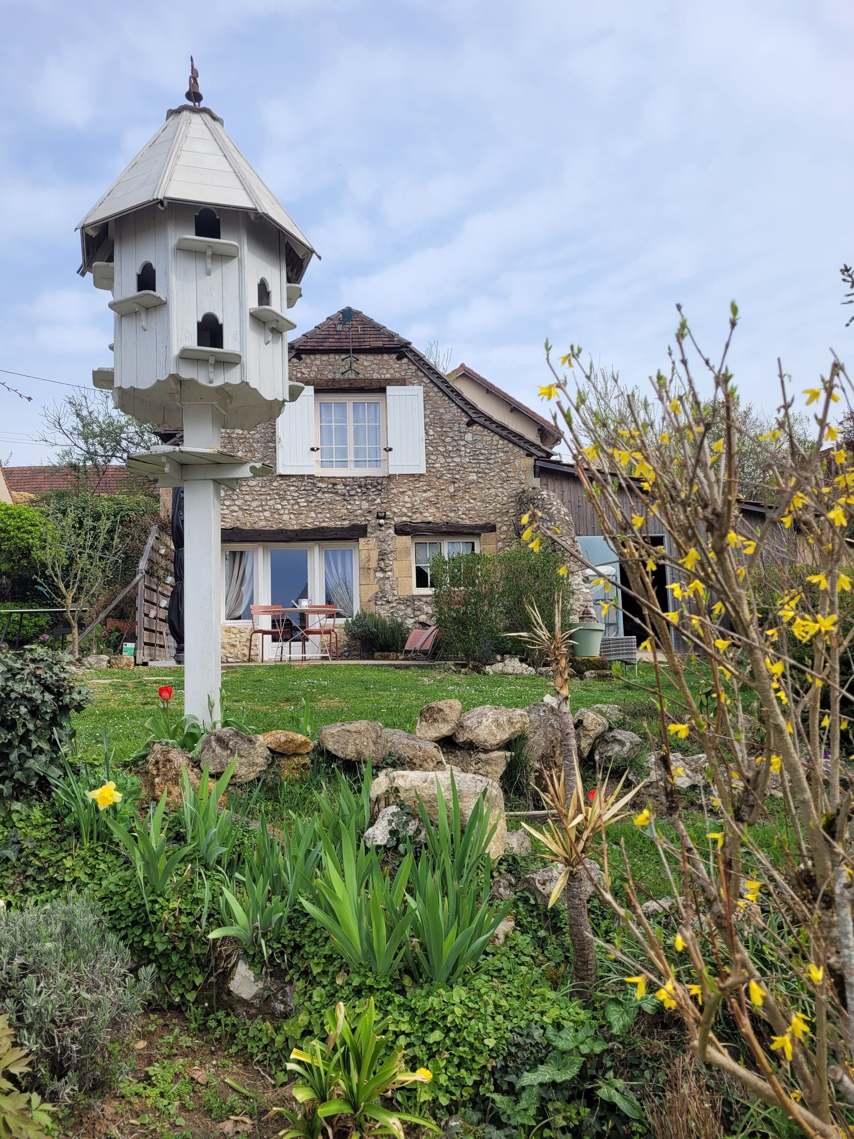 Ein Steinhaus mit einem Glockenturm, ein Garten mit Steinen und Pflanzen, und ein klarer Himmel.
