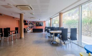 A spacious indoor area with multiple tables and chairs, large windows, and a colorful wall decoration.