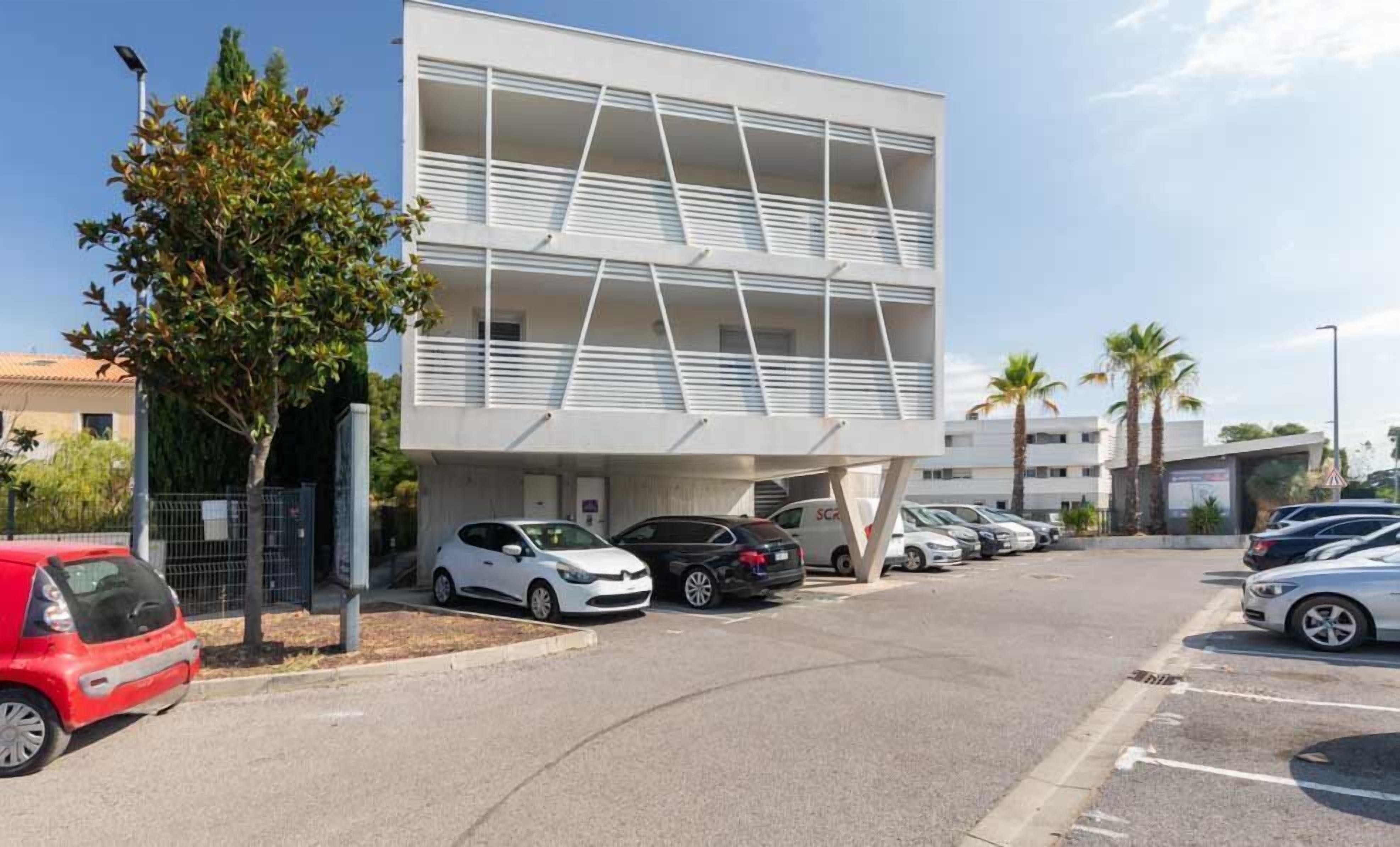 A modern building with a flat roof, multiple balconies, and a parking area with several cars.