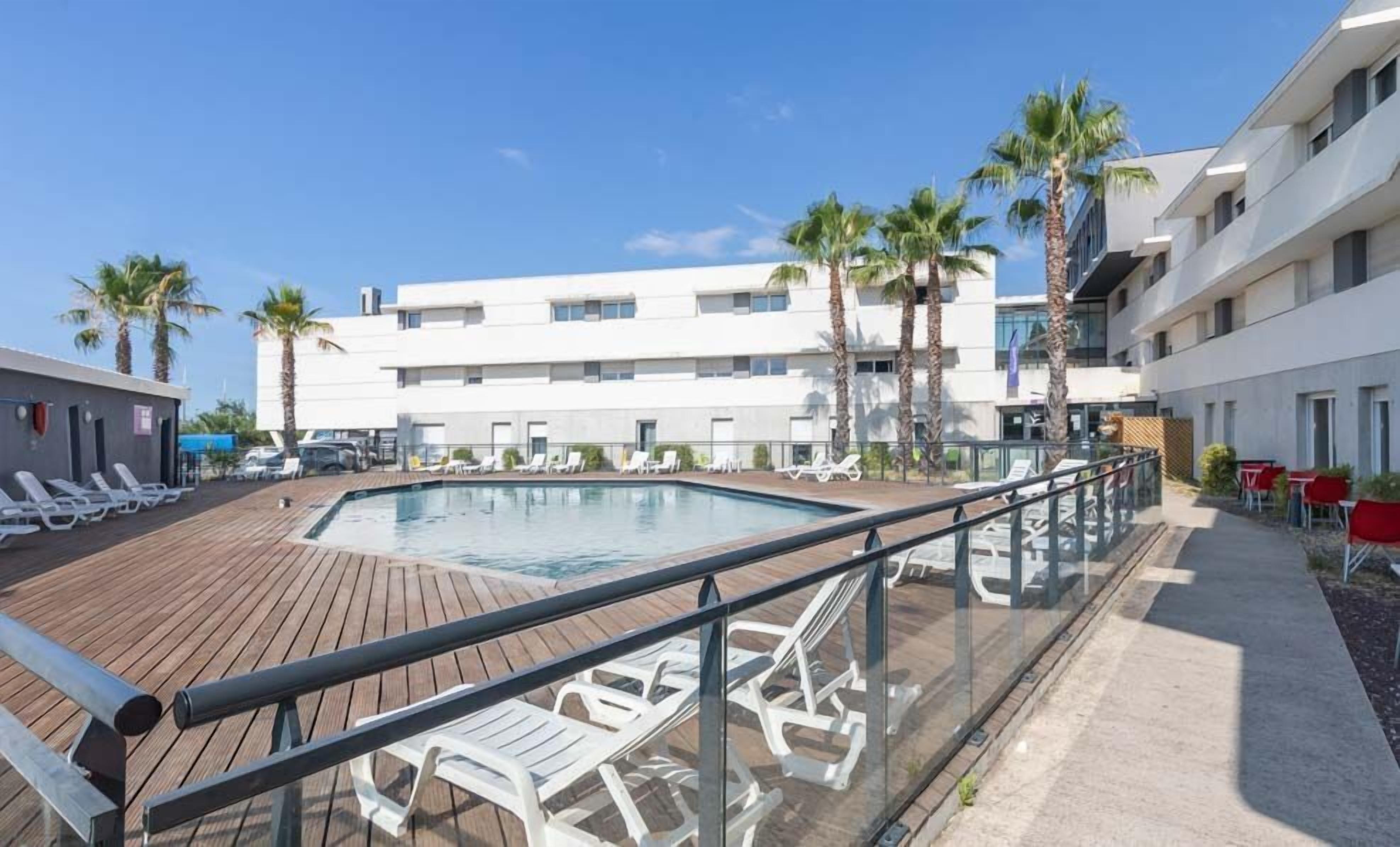 Un espace piscine d’hôtel avec une terrasse en bois, des chaises longues et des palmiers.