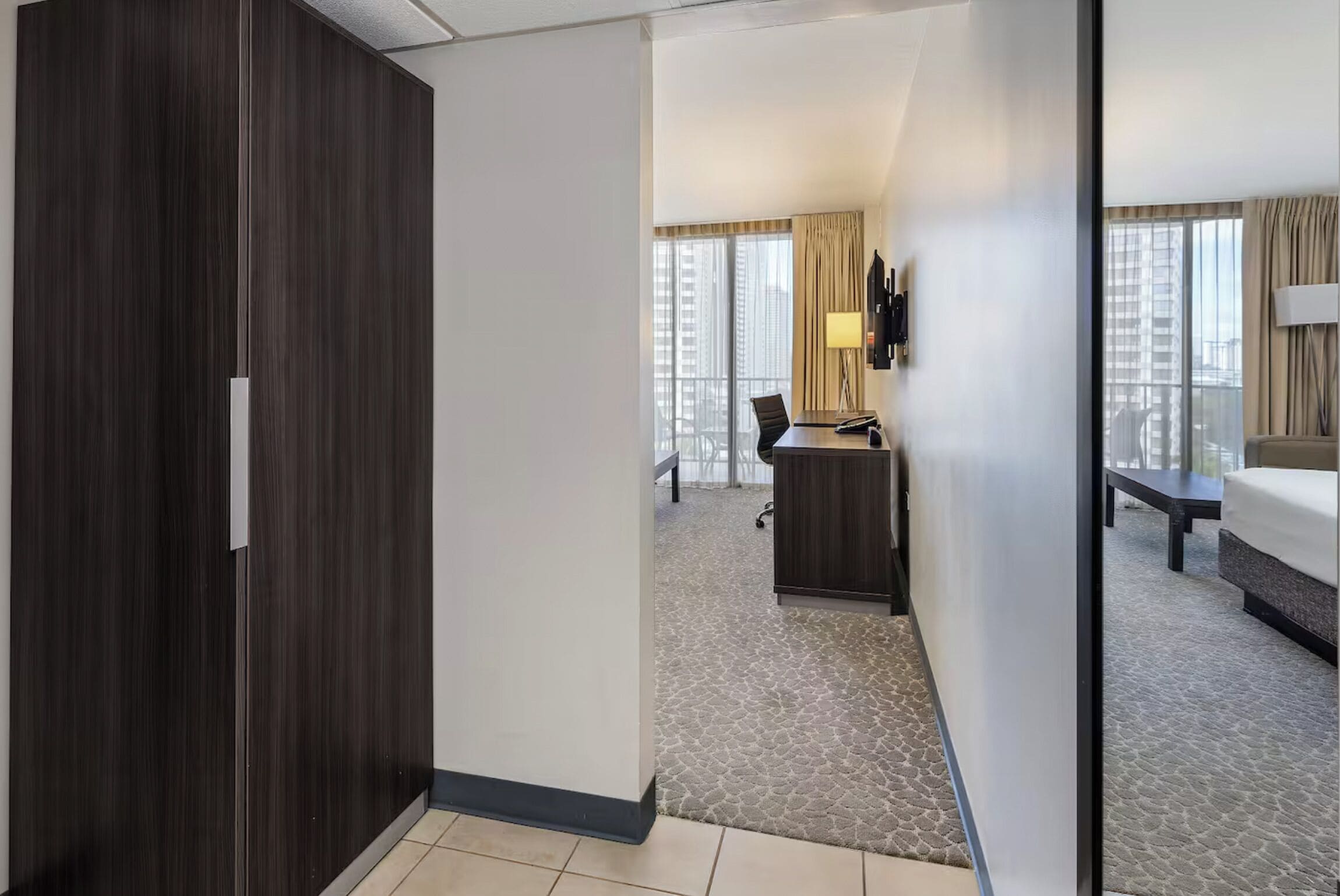 Quarto de hotel com guarda-roupa de madeira escura, uma escrivaninha com cadeira, uma cama e uma janela grande com cortinas.