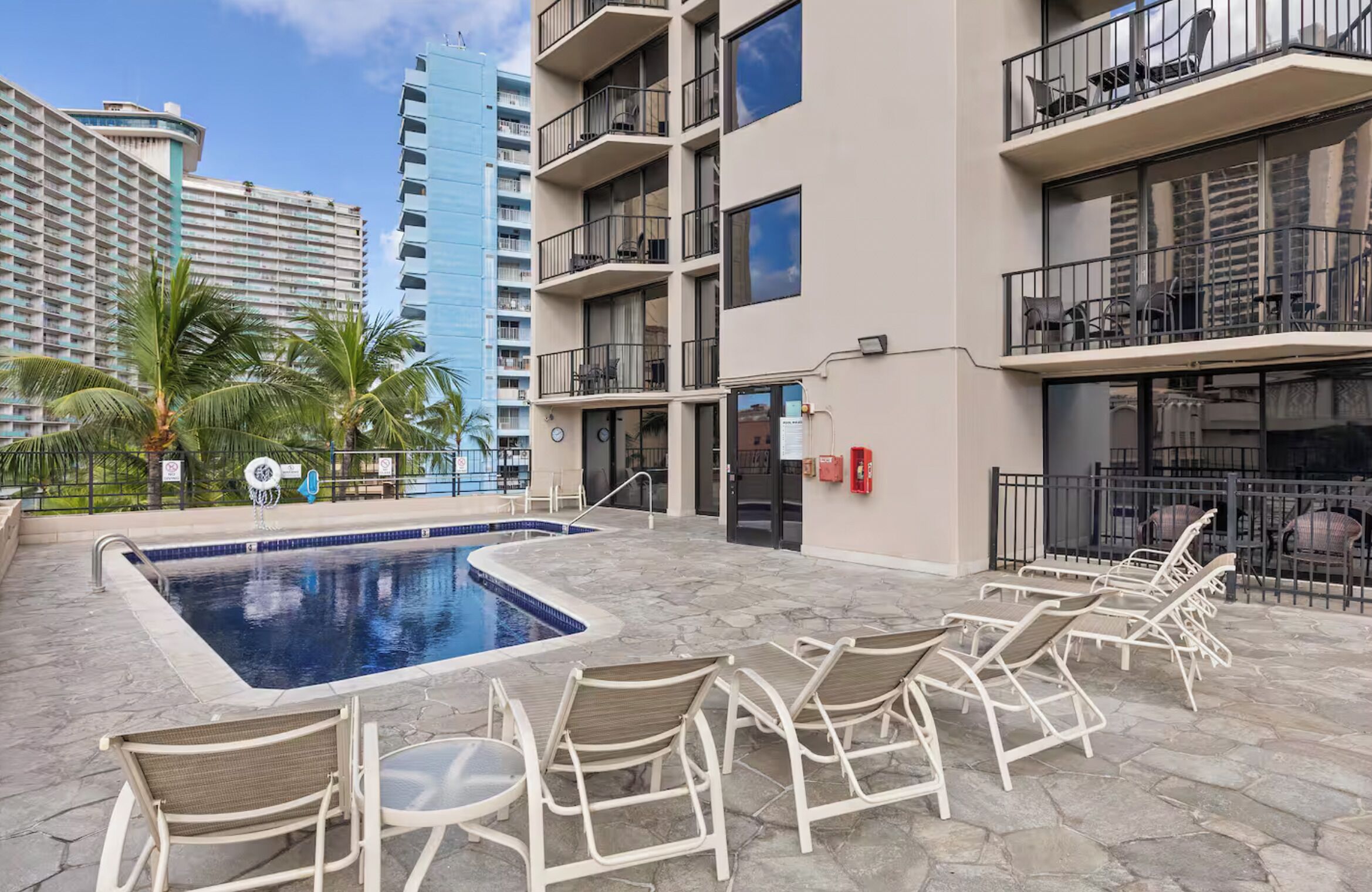 Un espace piscine avec des chaises longues, un bâtiment doté de balcons et un palmier.