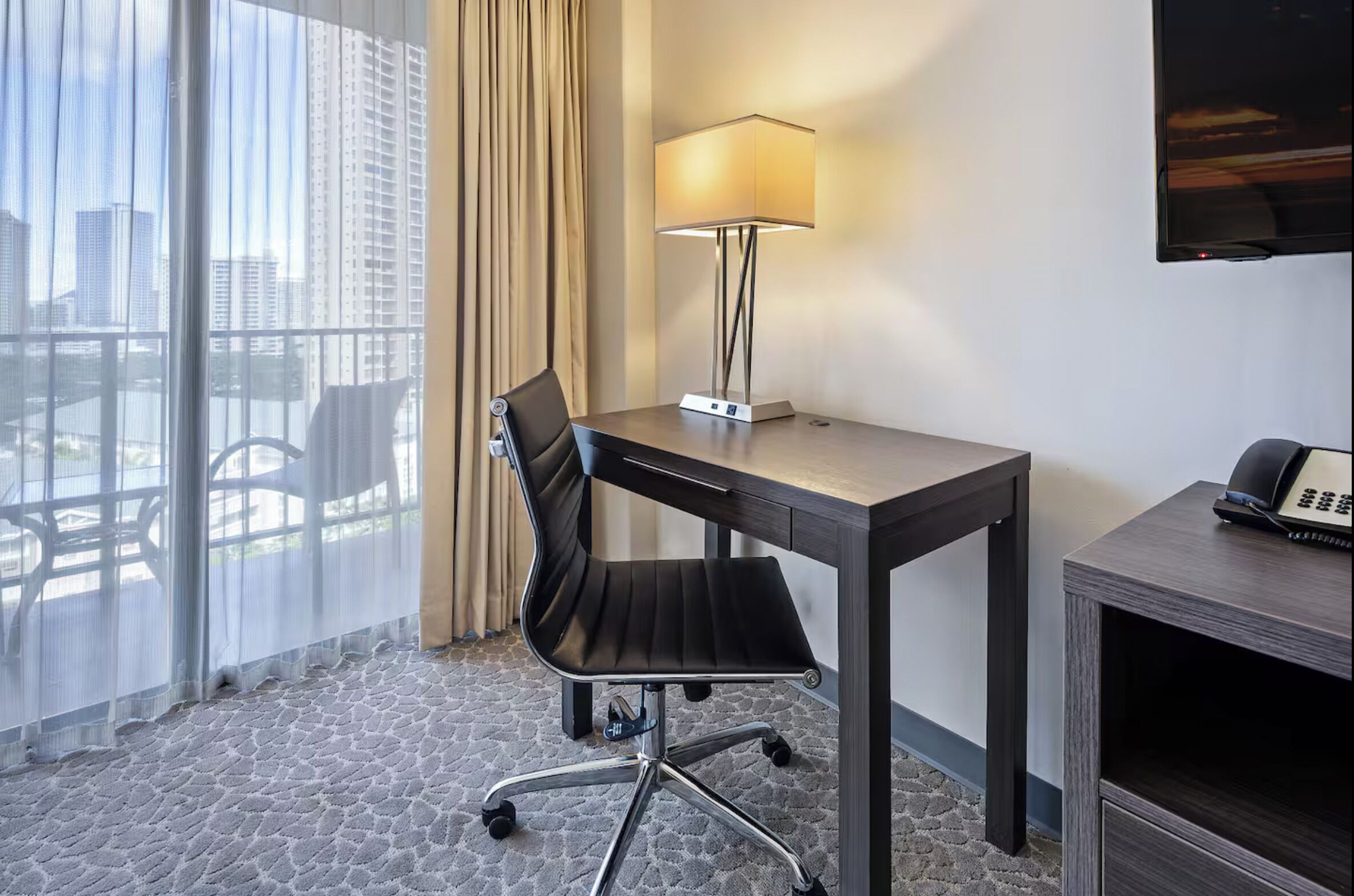 Una habitación de hotel con escritorio, silla, lámpara y teléfono sobre una mesita.