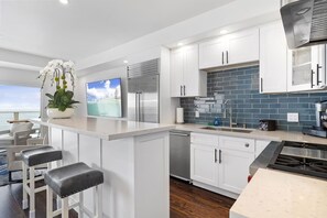 A modern kitchen with white cabinets, a stainless steel refrigerator, a dishwasher, and a blue-tiled backsplash. There are two bar stools and a potted plant on the counter.