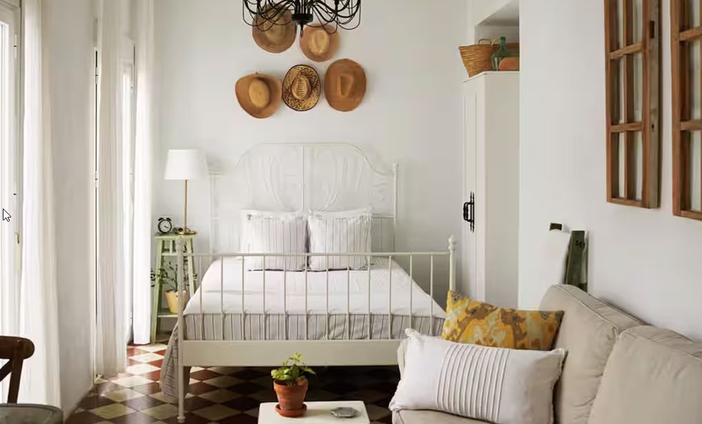 A bedroom with a white bed, a sofa, a checkered floor, and a wall with hats.