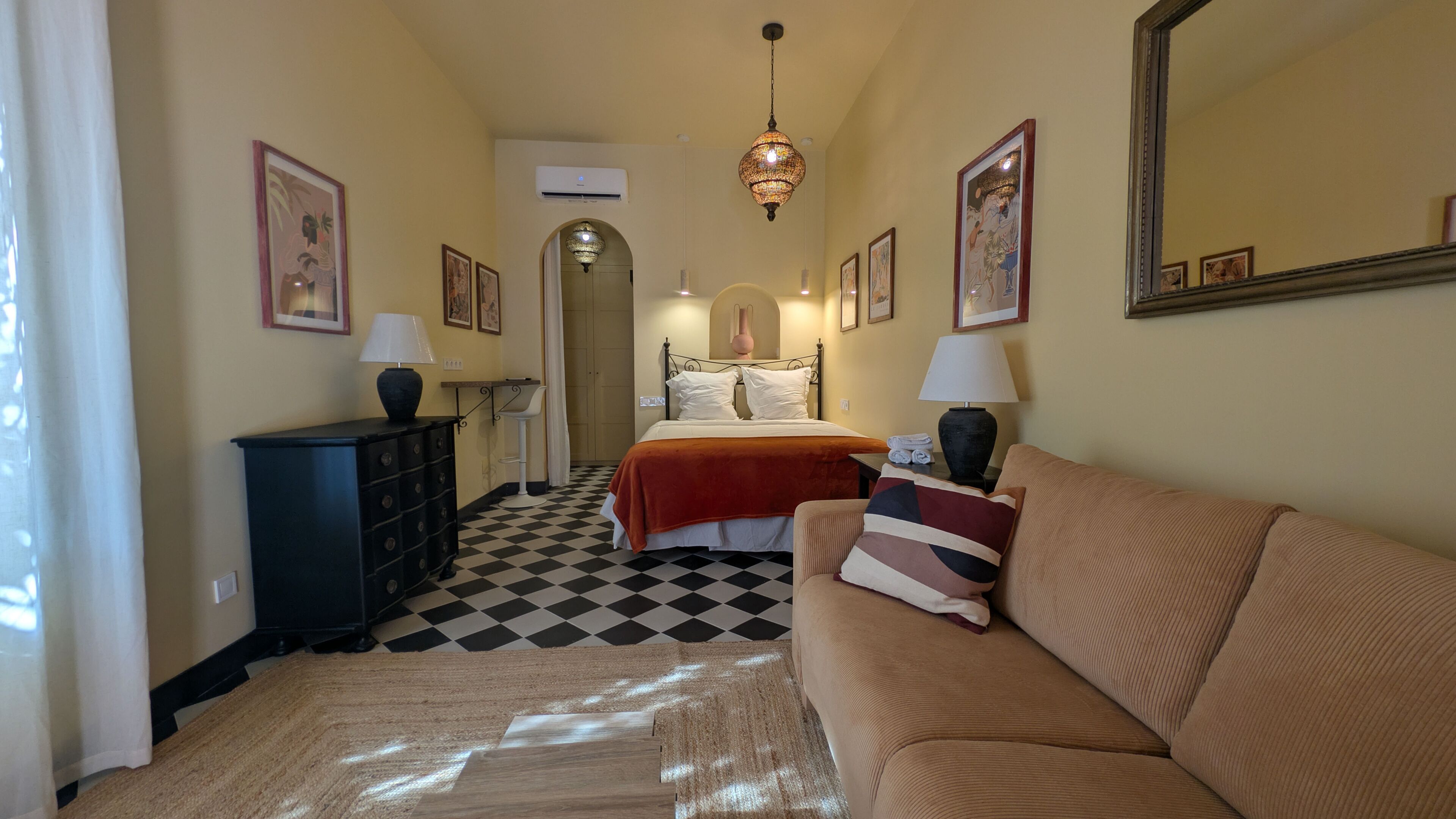 Habitación de hotel con piso a cuadros, una cama con ropa blanca, un sofá color beige y un aparador con lámparas.