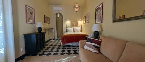 Habitación de hotel con piso a cuadros, una cama con ropa blanca, un sofá color beige y un aparador con lámparas.