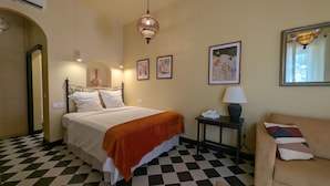 Ein Hotelzimmer mit Schachbrettmuster-Bodenbelag, einem Bett mit weißer Bettwäsche und einer orangefarbenen Decke, einem Nachttisch mit Lampe sowie einem Bild an der Wand.
