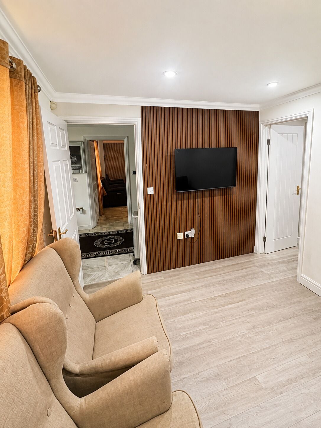 Una sala de estar con un sofá color beige, un televisor de pantalla plana montado en una pared de paneles de madera, y una puerta que da a otra habitación.