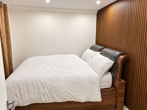 A bed with a white floral-patterned bedspread and matching pillows, a wooden headboard, and a wooden panel wall. - One Double Bedroom furnished plus living room Self Contain with Shared Kitchen (Orpington)