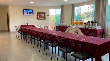 Sala de conferência com uma mesa comprida, cadeiras, uma televisão e um buffet com bebidas.