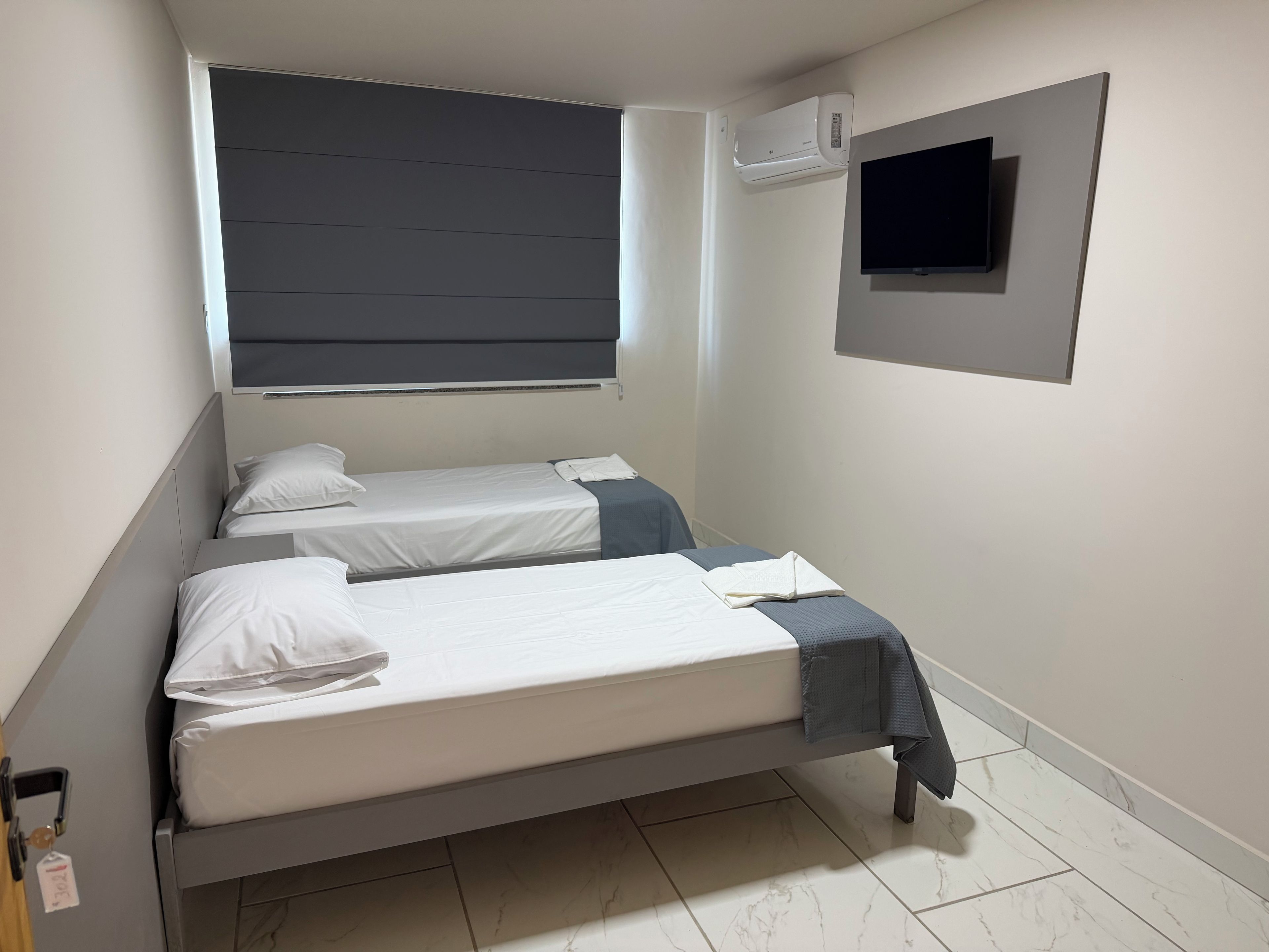 Quarto pequeno e moderno, com cama, janela com persianas, ar-condicionado fixado na parede e TV tela plana.