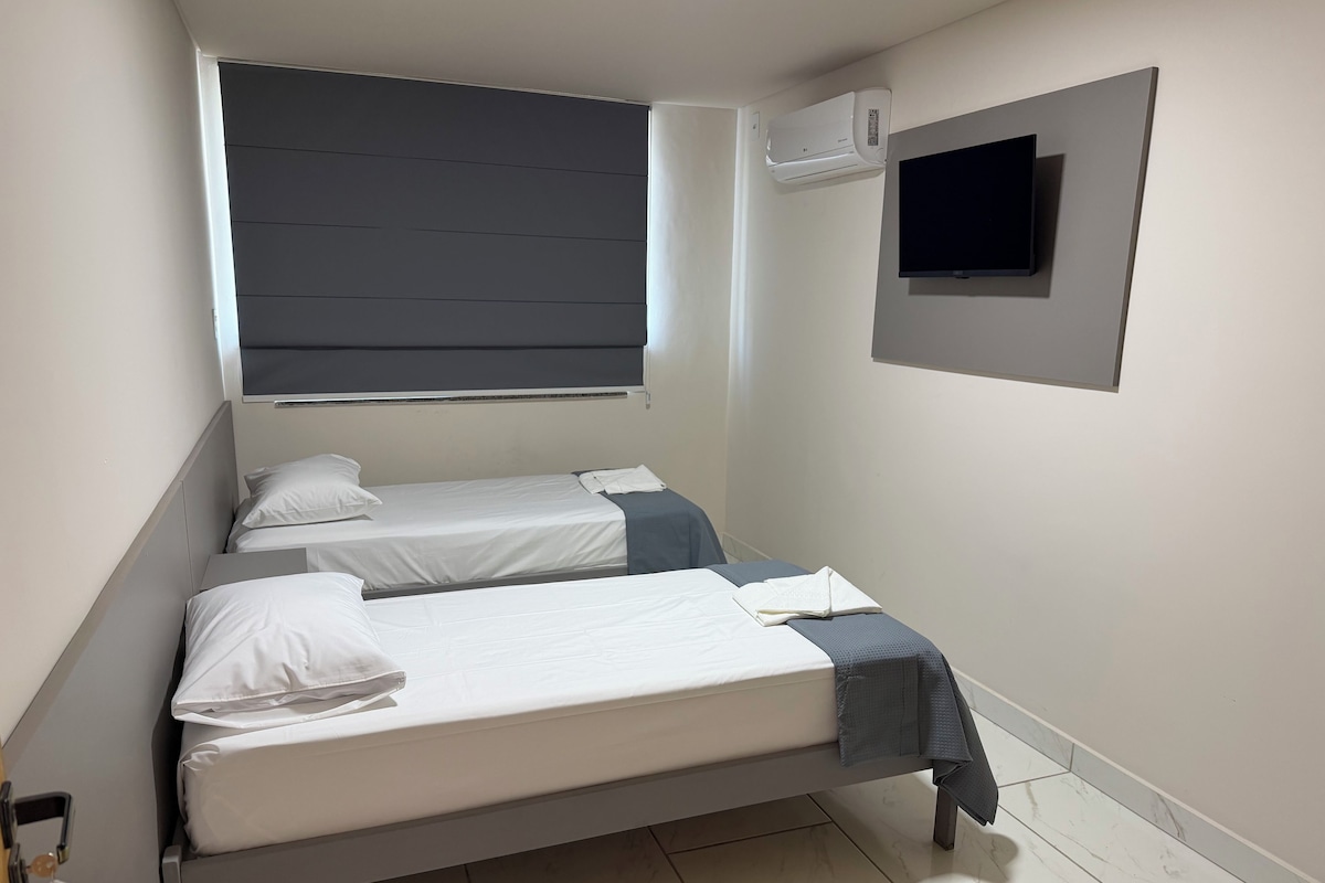 Una habitación pequeña y moderna con una cama, un ventanal con persianas, aire acondicionado de pared y una televisión de pantalla plana.