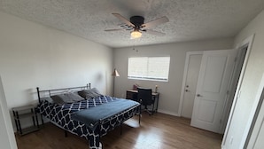 A bedroom with a bed, a desk, a chair, a window with blinds, a ceiling fan, and a door. - Parkside Retreat - Central DFW - Near AT&T Stadium, DFW Airport & Arlington (Grand Prairie)