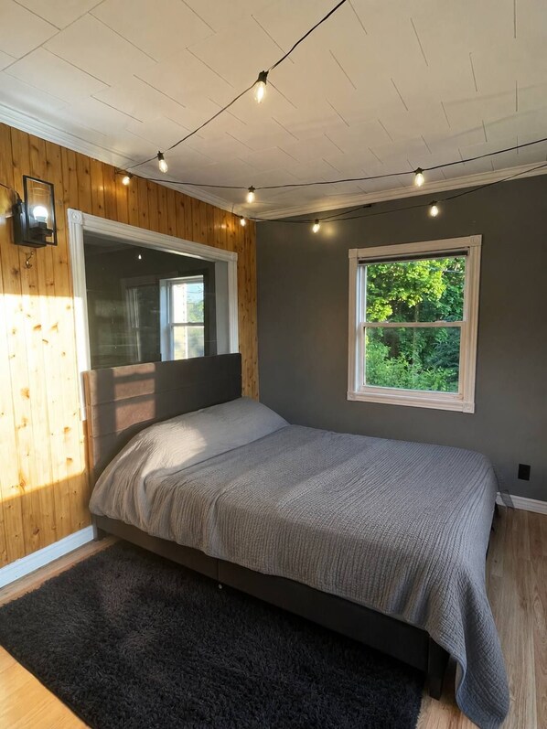 A bedroom with a bed, a window with a view of greenery, and a wooden wall.