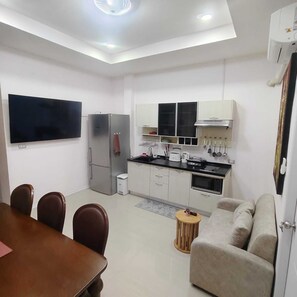 A modern kitchen with a flat-screen TV, a refrigerator, and a microwave. There is a dining table with chairs and a sofa in the living area. -  2-bedroom luxurious house with swimming pool. (Tambon San Sai Noi)