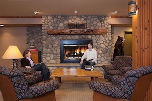 A cozy lounge area with a stone wall, a fireplace, patterned furniture, and a wooden table.