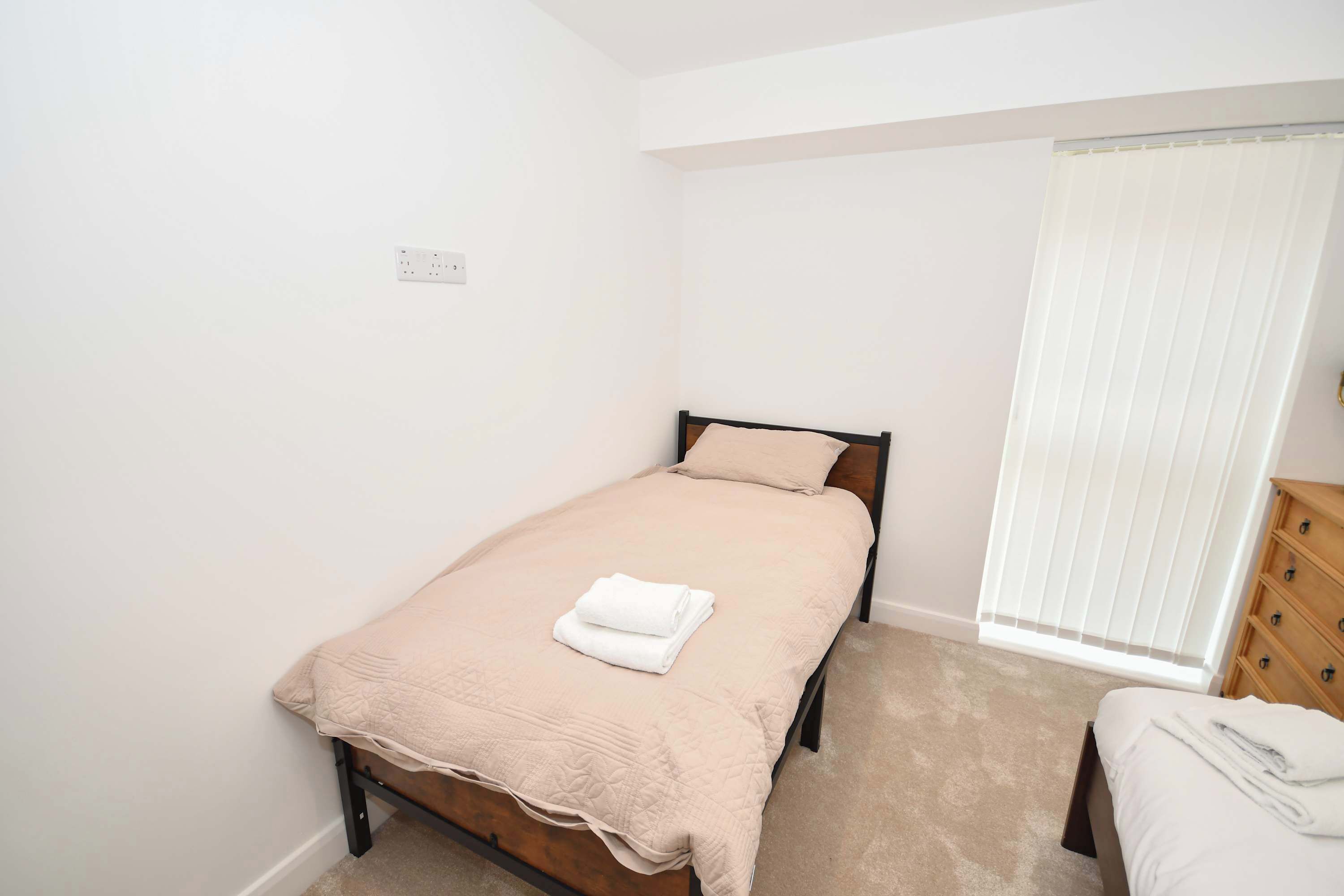 A single bed with a beige quilt and a white towel, a wooden nightstand, and a window with blinds.