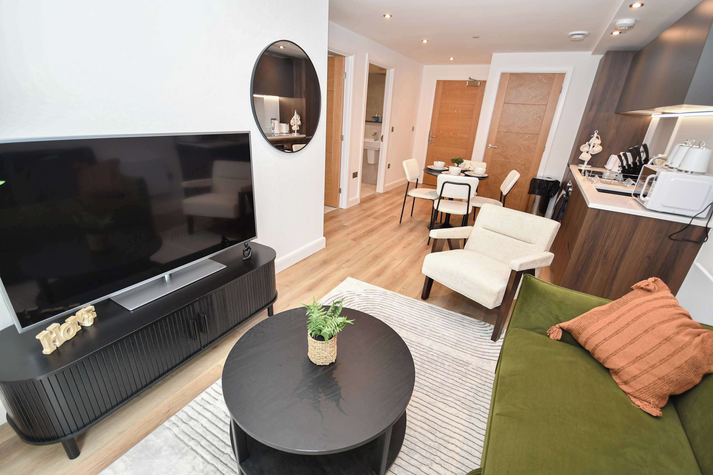A modern living room with a large flat-screen TV, a black coffee table, a green sofa, and a dining area with a wooden table and chairs.