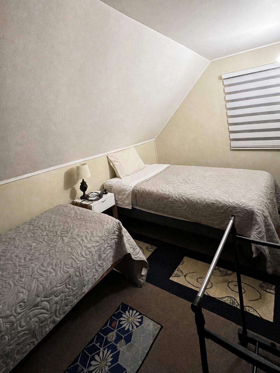 Una habitación pequeña y funcional con dos camas individuales, una mesita de noche con lámpara y una ventana con persianas.