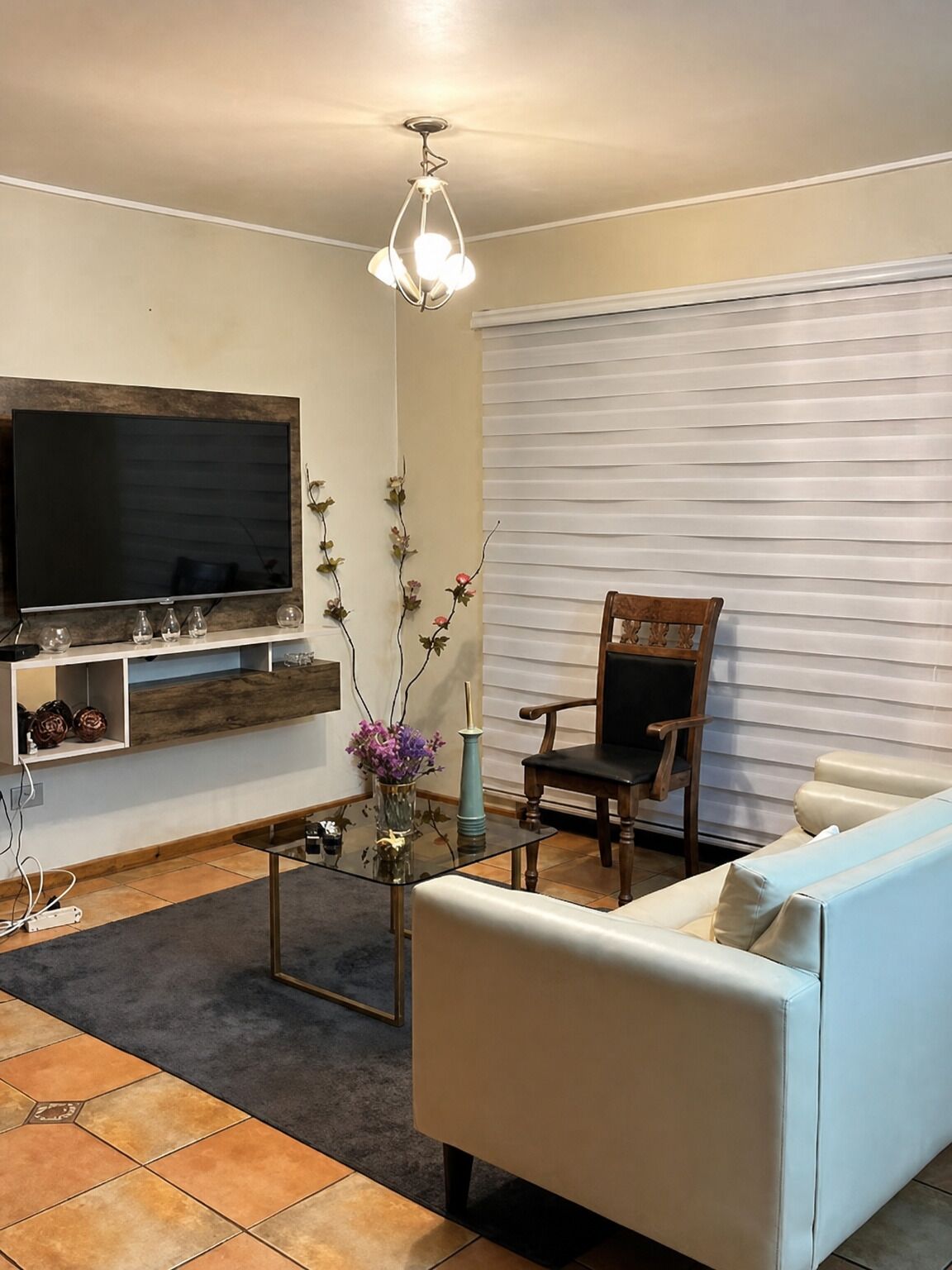 A living room with a flat-screen TV, a wooden entertainment unit, a glass coffee table, a white sofa, a wooden chair, and a window with blinds.