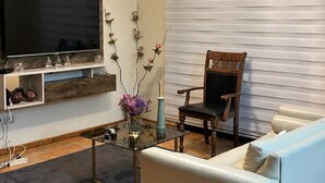 A living room with a flat-screen TV, a wooden entertainment unit, a glass coffee table, a white sofa, a wooden chair, and a window with blinds. - Cozy and spacious home with parking (Los Angeles)