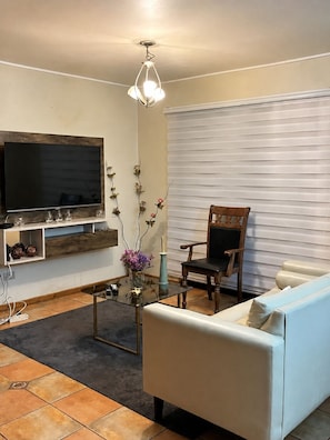 Una sala de estar con televisión de pantalla plana, un mueble de entretenimiento de madera, una mesa de centro de vidrio, un sofá blanco, una silla de madera y una ventana con persianas.