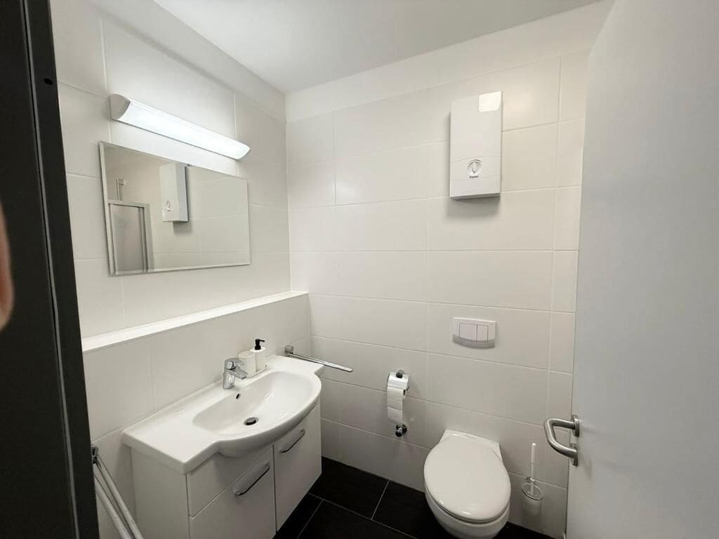 Un baño moderno con lavamanos blanco, espejo, inodoro y un calentador mural.
