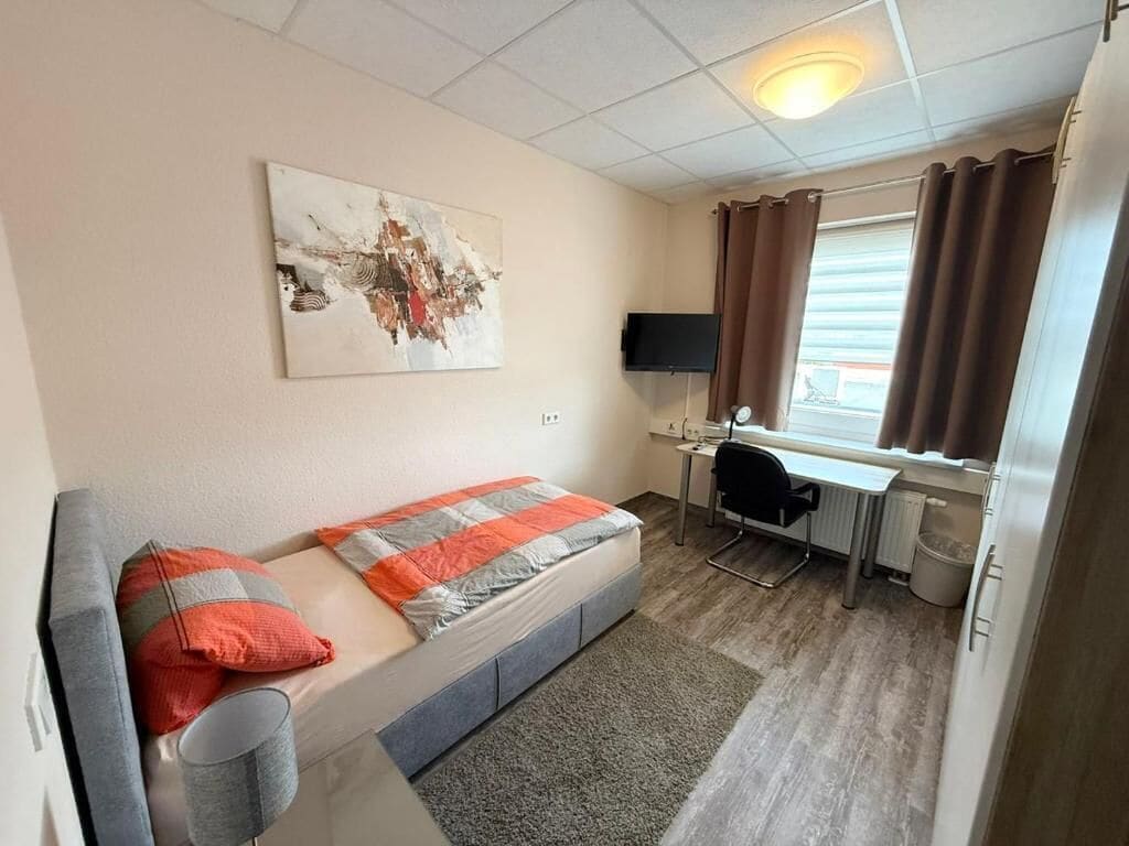 A compact room with a bed, a desk, a chair, a television, and a window with curtains.
