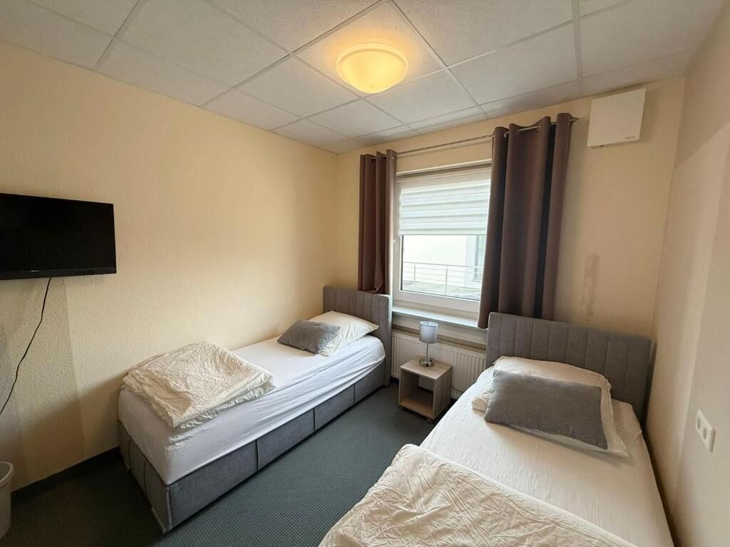 Una habitación de hotel con dos camas individuales, un televisor de pantalla plana montado en la pared, una ventana con cortinas y una mesita de noche con lámpara.