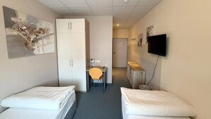 Quarto de hotel com duas camas de solteiro, uma escrivaninha com cadeira, um guarda-roupa, televisão presa na parede e uma porta que dá para outro quarto.