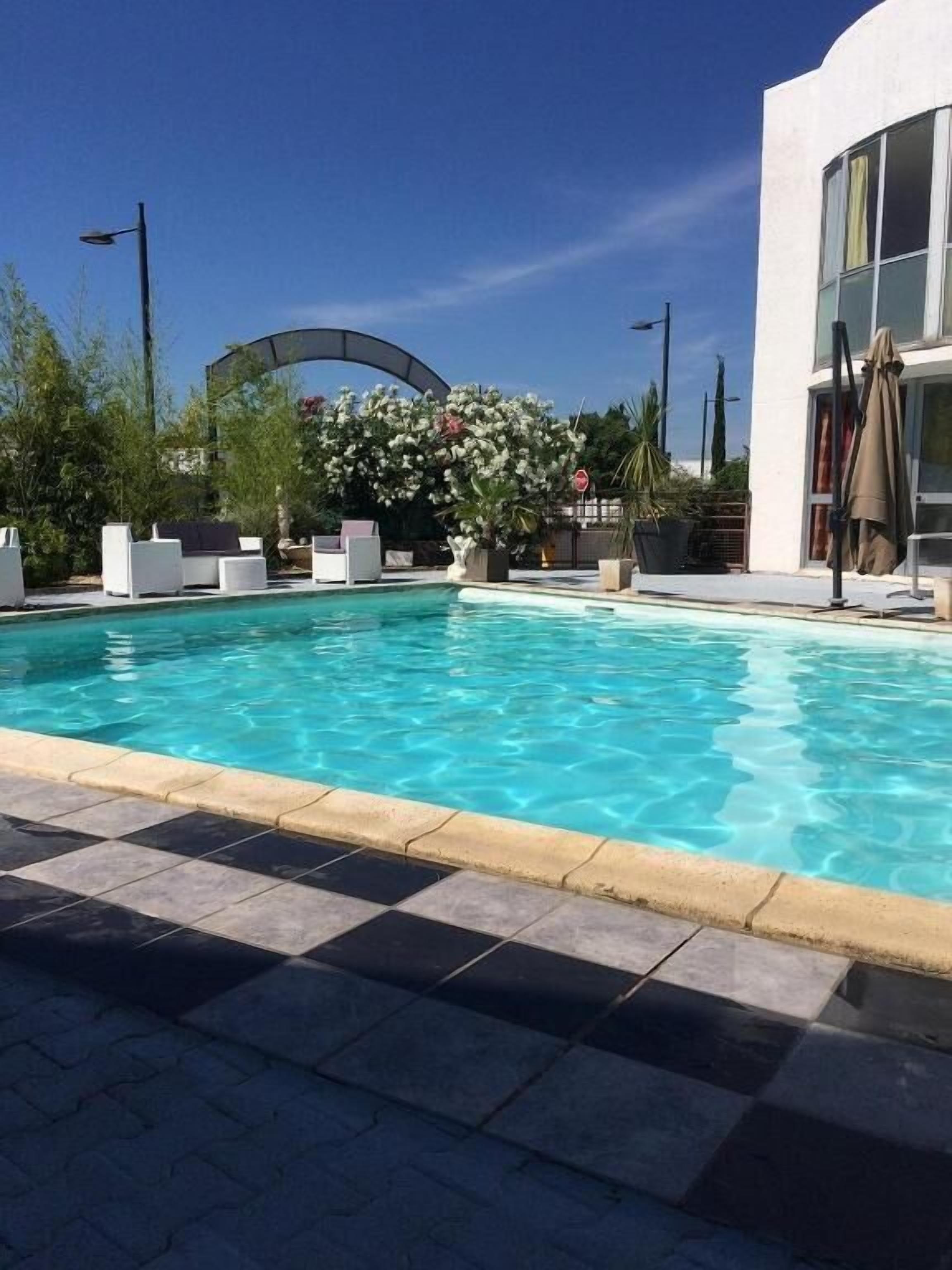Una piscina con un borde adoquinado, sillas de descanso y un edificio con grandes ventanales.