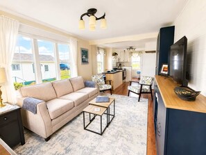Ein modernes Wohnzimmer mit Sofa, Couchtisch und Sesseln, eine Küchenzeile mit Esstisch und ein an der Wand befestigter Fernseher.