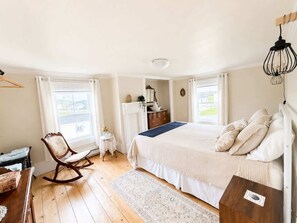 Ein Schlafzimmer mit einem großen Bett, einem Schaukelstuhl, einem kleinen Tisch und Blick ins Freie durch ein Fenster.