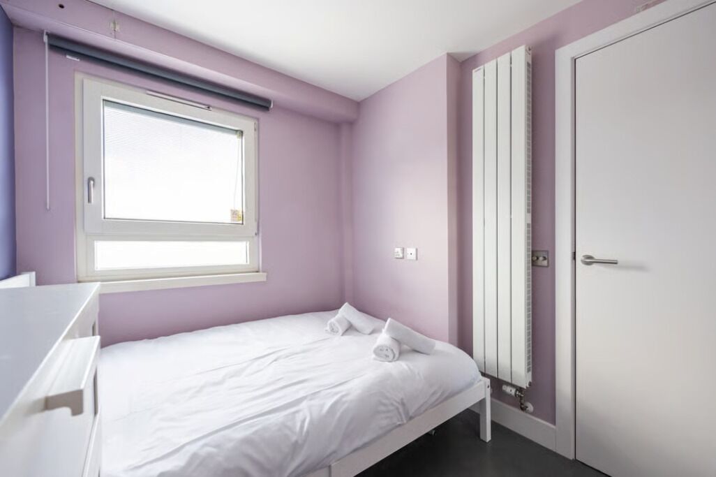 A bedroom with a single bed, a window with blinds, a radiator, and a door.