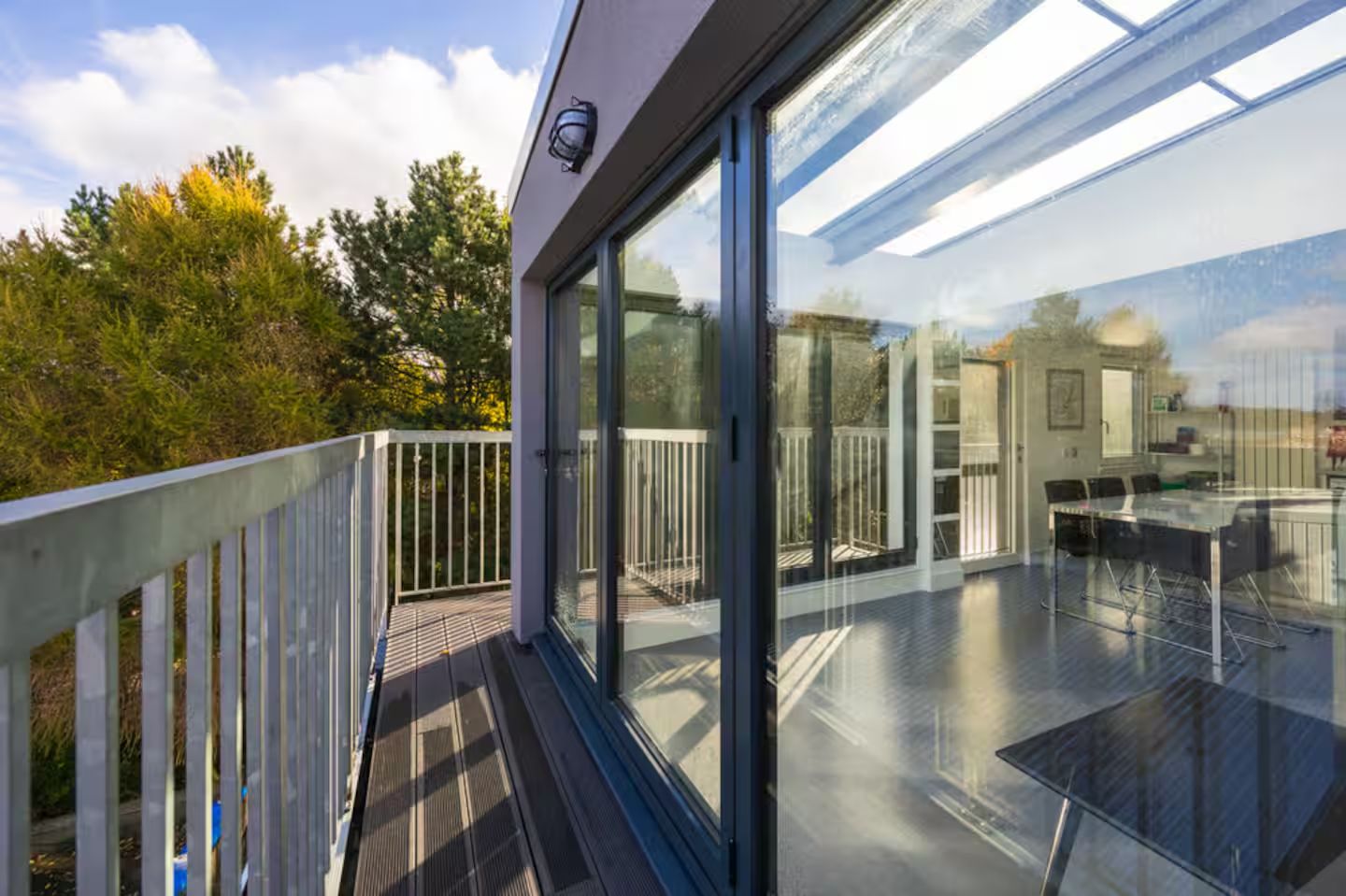 A modern building with large glass windows, a balcony with a white railing, and a view of trees and a clear sky.