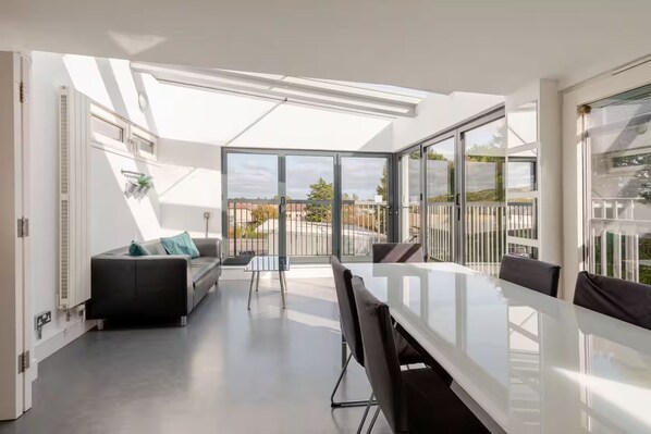 Un interior moderno con una mesa de comedor de cristal, sillas negras, un sofá con cojines y vistas amplias por grandes ventanales.
