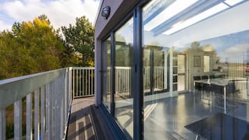 A modern glass-enclosed balcony with a wooden deck, a glass table, and a view of the surrounding trees.