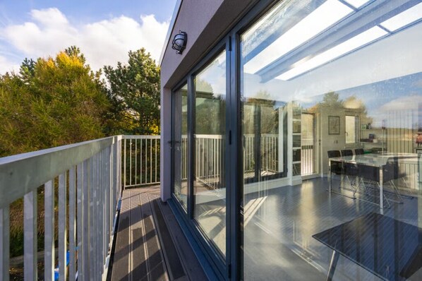 A modern glass-enclosed balcony with a wooden deck, a glass table, and a view of the surrounding trees.