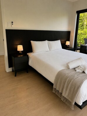 A modern bedroom with a large bed, two bedside tables with lamps, a window with a view of greenery, and a black headboard. - The NÖRA • Luxury Forest Retreat, Hot Tub • Sauna (La Conception)