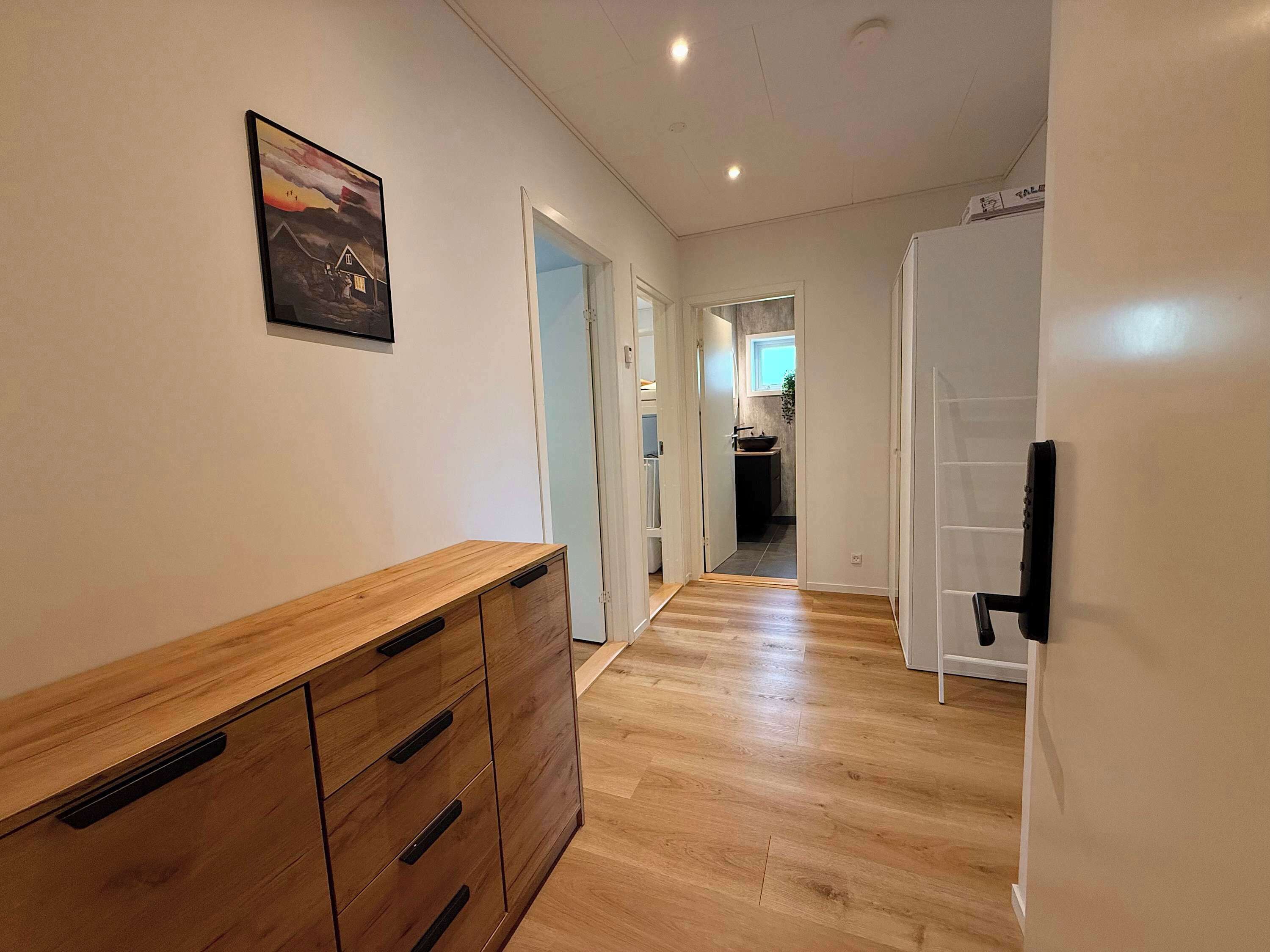 Ein Flur mit Holzboden, einem Holzkabinett mit Schubladen, einem gerahmten Bild an der Wand und einer Tür, die in einen Küchenbereich führt.