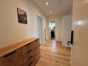 A hallway with wooden flooring, a wooden cabinet with drawers, a framed picture on the wall, and a door leading to a kitchen area.