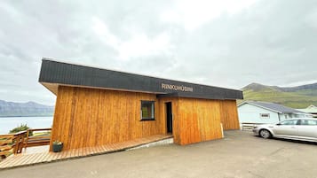 Une cabane en bois avec un toit noir, portant l’inscription « RINKUHUSINI », située au bord d’un plan d’eau, avec des montagnes en arrière-plan.
